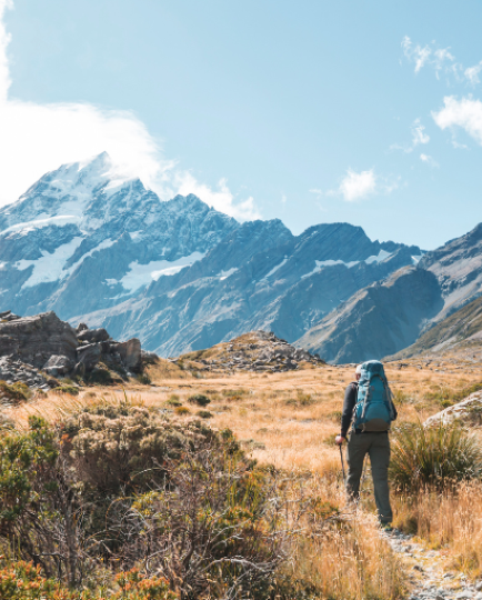 A man walks up a path, which leads to a mountain in the background. He is wearing a backpack suggesting he is tramping