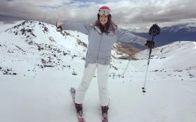 Katie standing on mountain in ski gear with arms out stretched looking happy