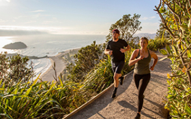 Two people run along a trail with the Tauranga harbour in the background