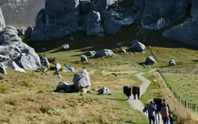 A group of people walk down a dirt road towards some rocky hills