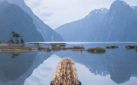 A lady stands in front of a lake, facing two mountains. The location is Milford Sounds in New Zealand.