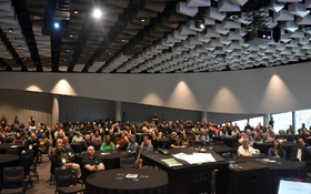 A large group of people gather in a conference room, they are sitting at round tables