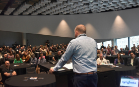 A man stands on a stage delivering a speech to the crowd. Photo is taken from behind him.