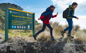 Two older children in hiking shoes and backpacks are walking past a Department of Conservation sign titled "Mangawhai Cliffs Walkway." The ground they are on is sandy with beach/coastal grasses growing on it. In the background is a blue sky and a small hill covered by trees.